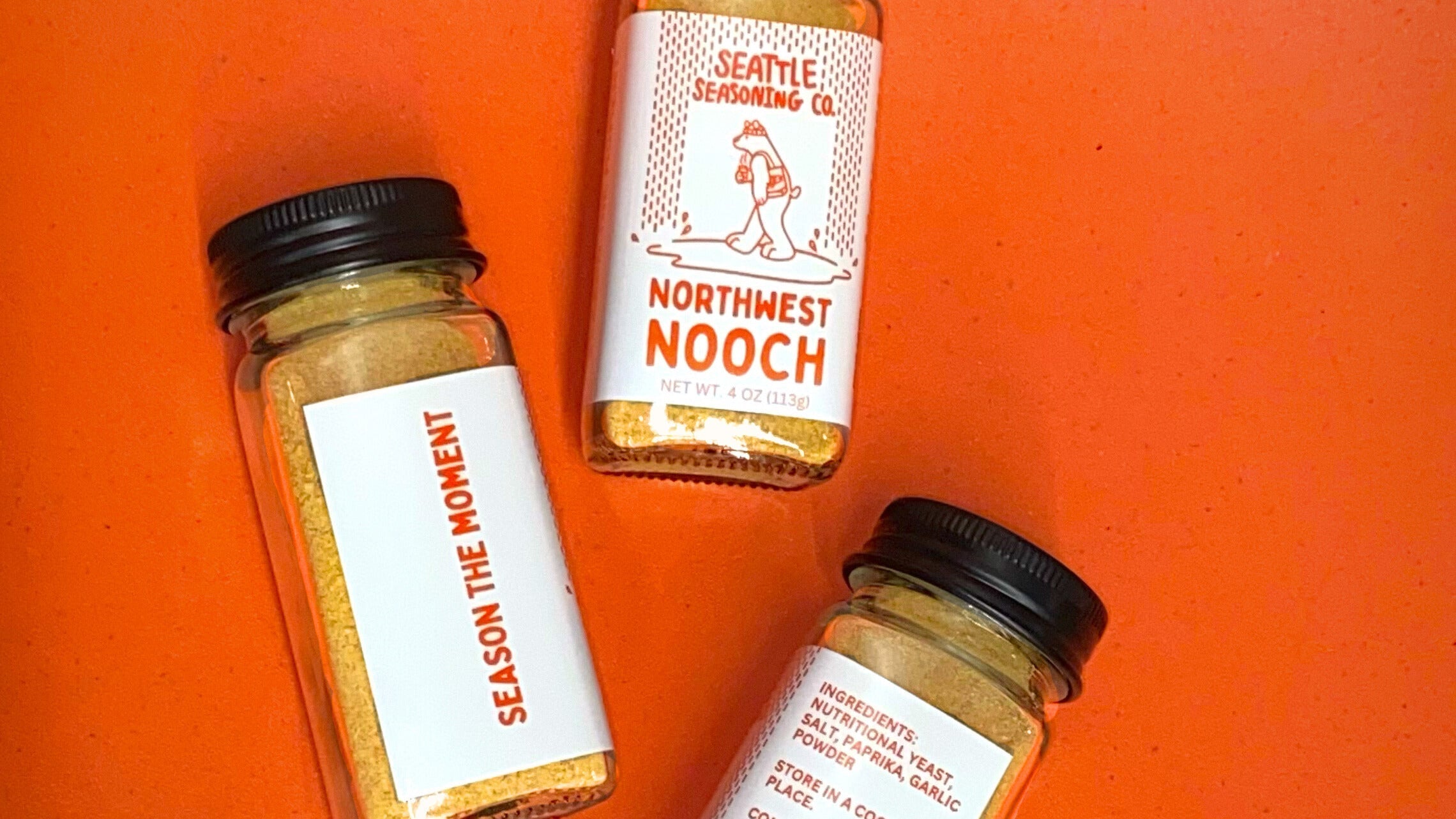 Three glass bottles of nutritional yeast seasoning labeled "Seattle Seasoning Co. Northwest Nooch".